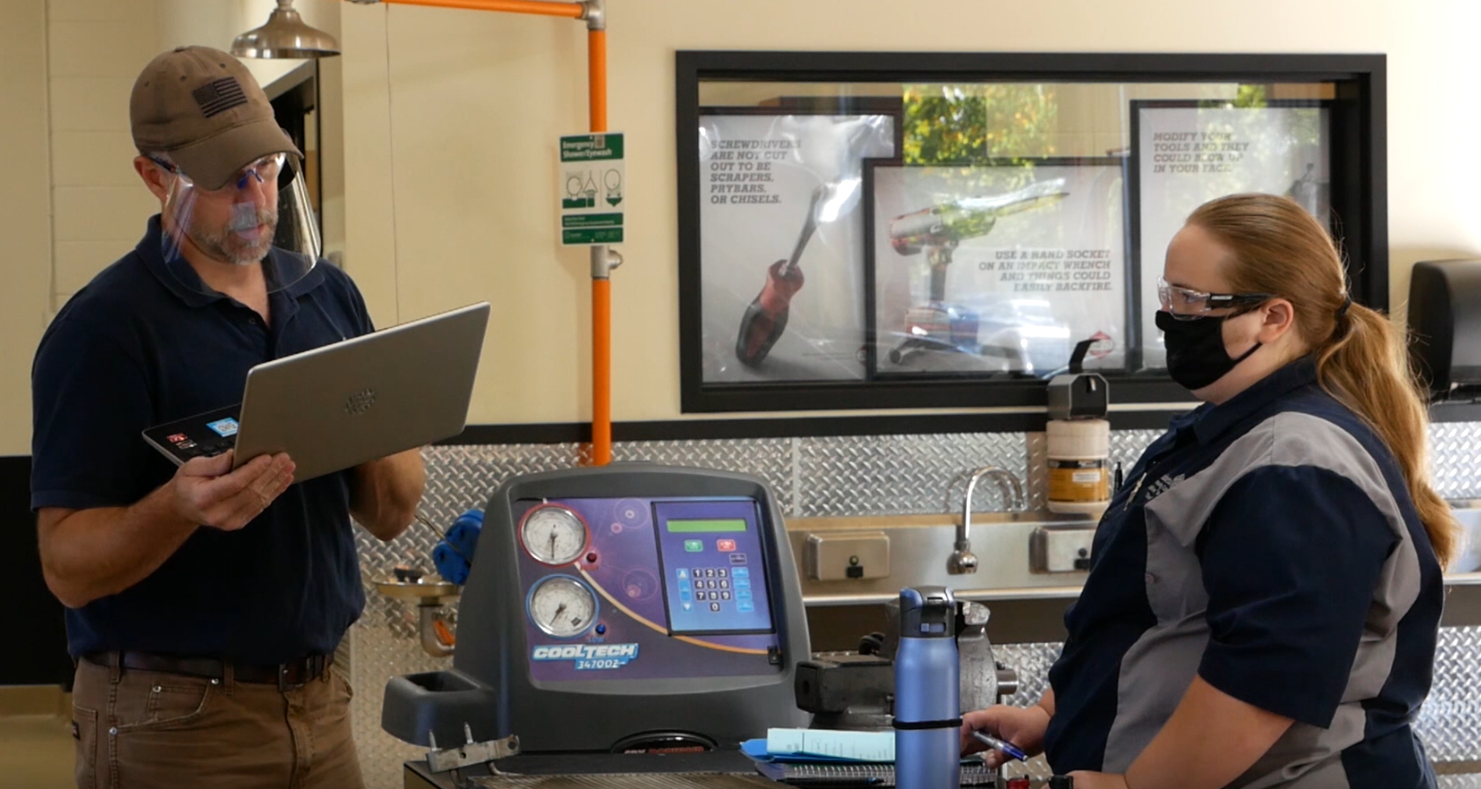 A student with a facemask and instructor with a face shield communicate during an automotive lab